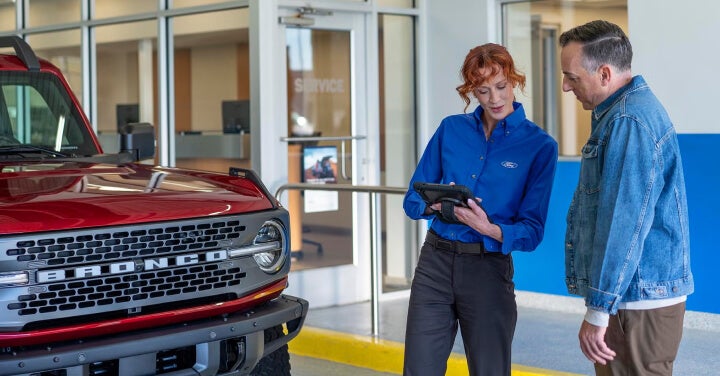 Ford advisor shows a tablet to a customer beside a red Bronco while discussing quality used vehicles at the dealership.